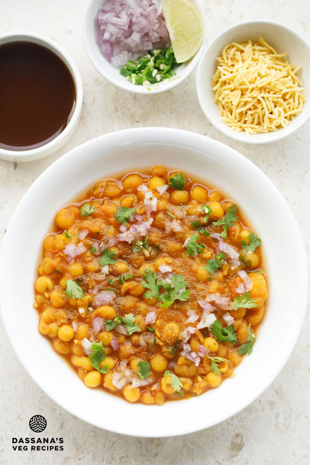 ghugni in white bowl topped with finely chopped onions, green chillies, coriander leaves with three side bowls on top filled with tamarind water in first bowl, onions, chillies and lemon wedges in second bowl and sev in the third bowl.