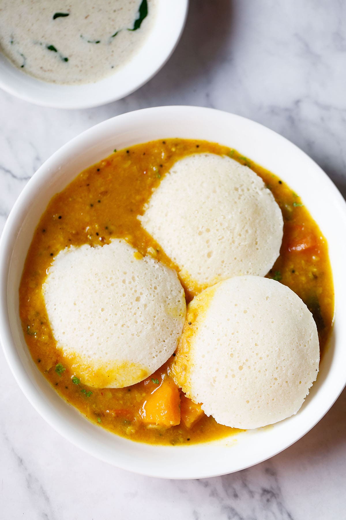 three idli on top of a layer of sambar in a white bowl with coconut chutney in a small bowl on top left.
