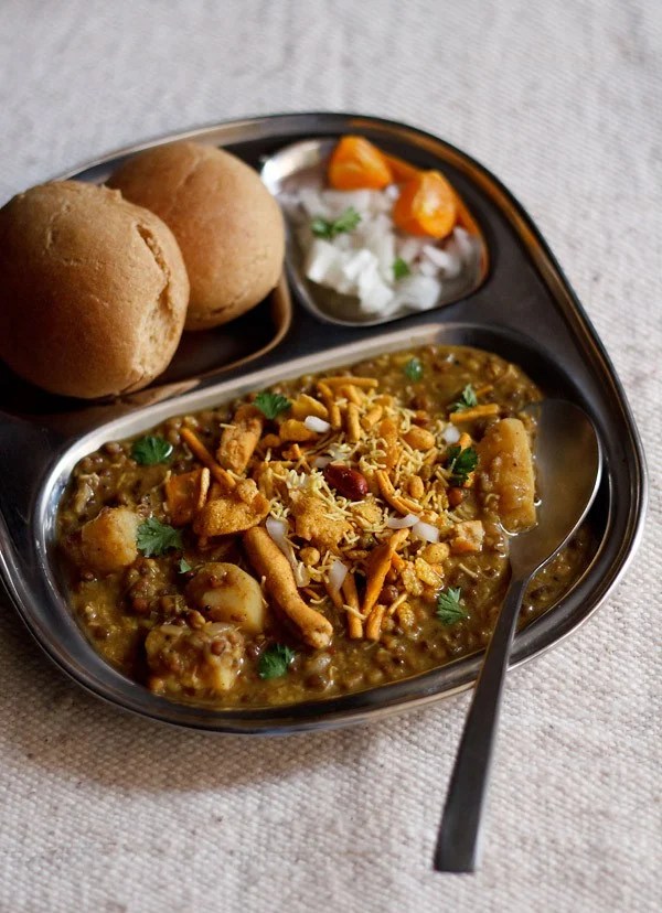 misal pav served in a plate topped with farsan with a side of sliced lemons and chopped onions and pav.