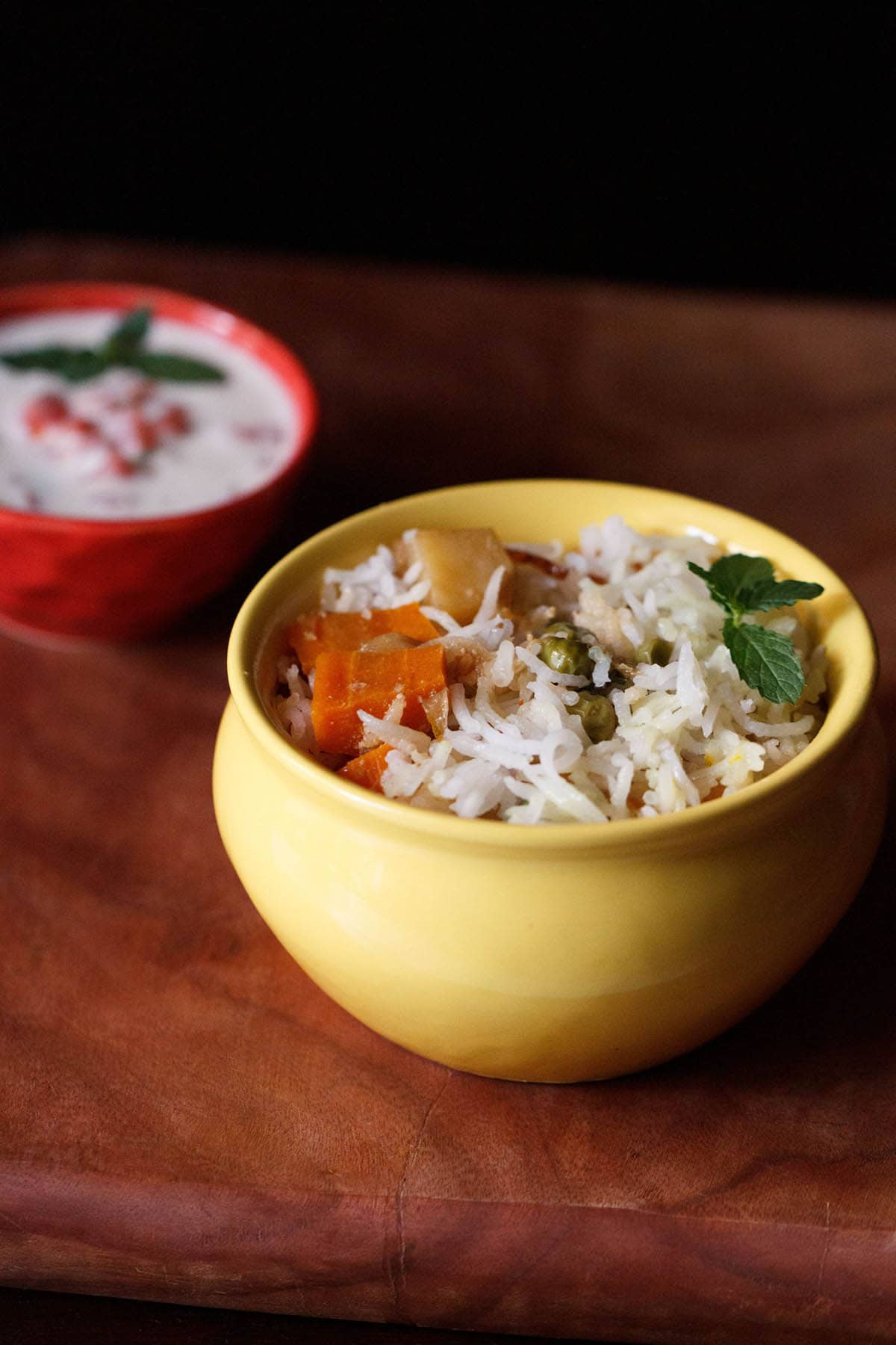 vegetable biryani served in a ceramic yellow handi with a bowl of raita on the side.